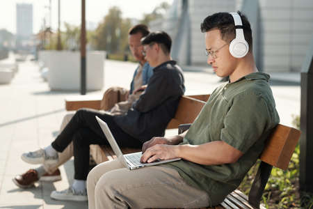 Young businessman in headphones and casualwear typing on laptop keypad while sitting on bench...