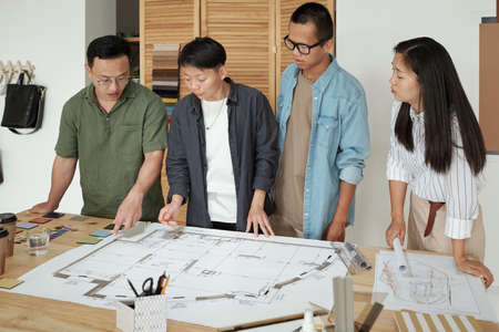 Group of young architects gathered by table with large sketch on blueprint discussing working points at meetingの写真素材