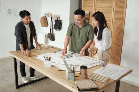 Three confident architects choosing color from palette on blueprint with sketch at working meeting while standing by tableの写真素材