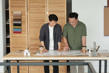 Two young contemporary engineers in casualwear standing by workplace and discussing sketch details while one of them pointing at blueprintの写真素材