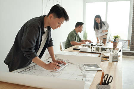 Young female architect or engineer drawing sketch while bending over table against her two co-workers in large openspace officeの写真素材