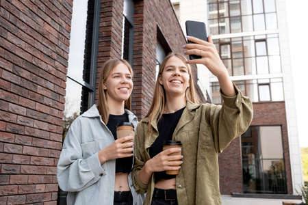 Smiling twin girls with drinks looking at smartphone camera while making selfie or communicating in...