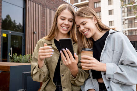 Young twin sisters in casualwear looking at smartphone screen while relaxing and having drinks in urban environmentの写真素材
