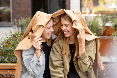 Two cheerful teenage girls covering heads with beige trenchcoat and looking one another while...