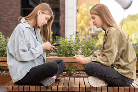 Twin girls sitting on bench in front of one another and scrolling in smartphones on summer day in urban environmentの写真素材