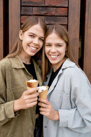 Beautiful twin sisters with long blond hair standing against wall of modern building and having icecream in front of cameraの写真素材