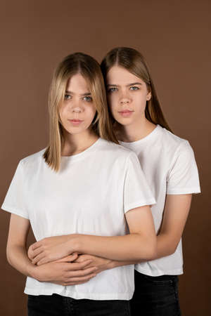One of affectionate twin girls in white t-shirts embracing her sister while both standing against brown backgroundの写真素材
