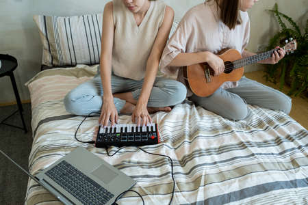 Two creative teenage girls in casualwear playing musical instruments and recording music while sitting on bedの写真素材