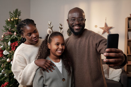 Happy young family of three making selfie by xmas tree while smiling man holding mobile phone in...