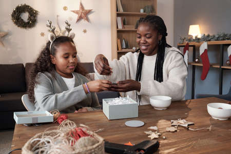 Cute African girl in headband helping her mother with packing of xmas gifts while sitting by table...