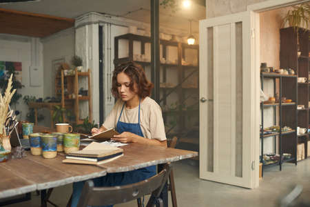 Young female ceramist in workwear making notes while sitting by table in front of laptop in her...