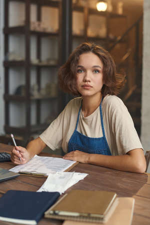 Young pretty female ceramist looking at camera while working with documents by table in her workshop