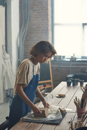 Young female master of pottery working with clay while standing by table in large contemporary...