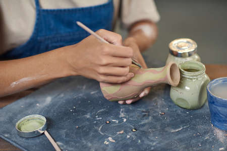 Hand of young female with paintbrush applying pink stripes on handmade jug or pot while sitting by...