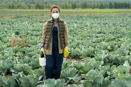 Full length portrait of female cabbage grower in respirator and gloves standing with sprayer on plantationの写真素材