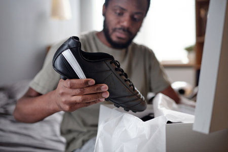 Hands of young African man with black sports-shoe putting it into bag or unpacking box with sporty...