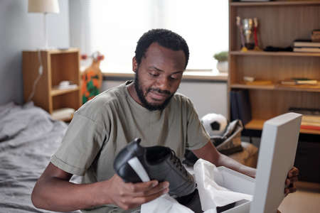 Young African sportsman unpacking box with new black sports-shoes while sitting on bed and preparing...