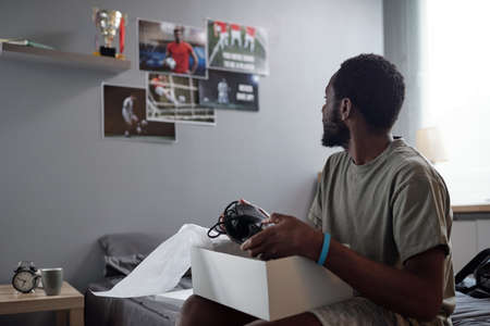 Sportsman of African ethnicity looking at posters of football players on wall while sitting on bed...