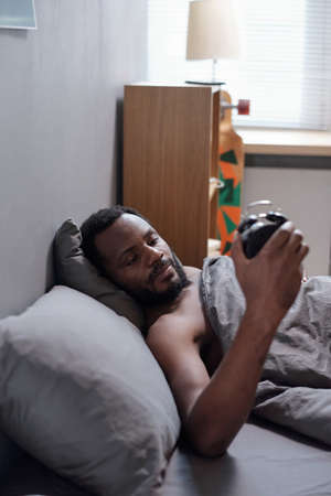 Young bearded man of African ethnicity lying under blanket in bed and looking at clock face after...