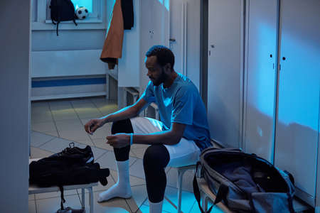 Young African soccer player going to put on his sports-shoes while sitting by row of lockers in...