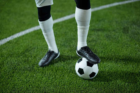Legs of contemporary young footballer with his foot on soccer ball standing on the field and waiting...
