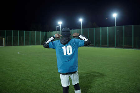 Back view of young football player putting forefingers in his ears while standing on green lawn at large contemporary stadiumの写真素材