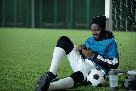 Young restful sportsman sitting by net and scrolling in smartphone while looking through news or online videosの写真素材