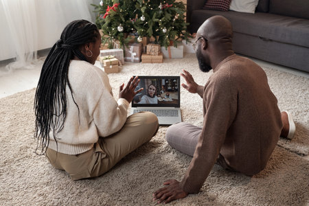 Young African couple communicating to their friend in front of laptop while sitting on the floor of living-room on Christmas dayの写真素材
