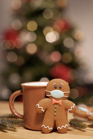 Conceptual gingerbread cookie with glazed protective mask standing by mug with hot drink on wooden tableの写真素材