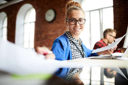 Happy young businesswoman working with papers while sitting by desk on background of colleagueの写真素材