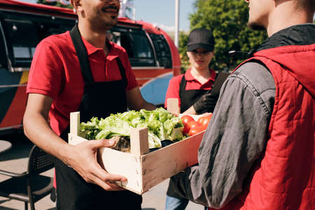 Young clerk in uniform passing wooden box with fresh tomatoes and lettuce to courierの写真素材