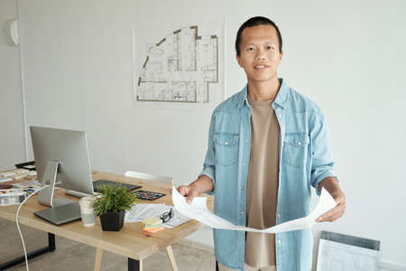Young Chinese creative designer with blueprint standing against his workplace