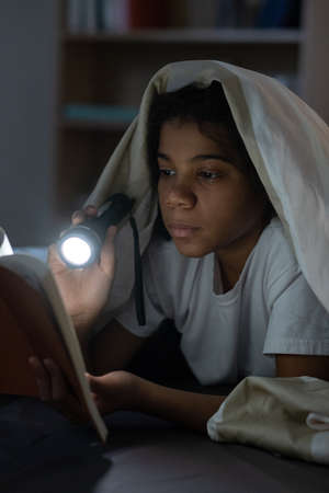 Concentrated Black girl lying under duvet and using flashlight while reading fairy tale in darkness