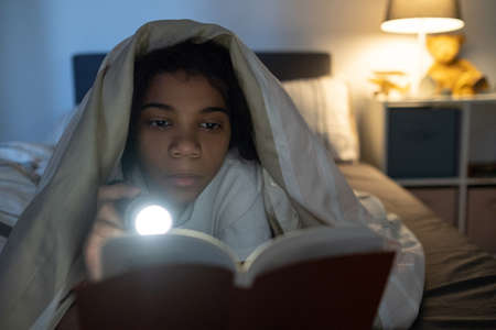 Serious African American girl lying under duvet on bed and shining flashlight on book in darknessの写真素材
