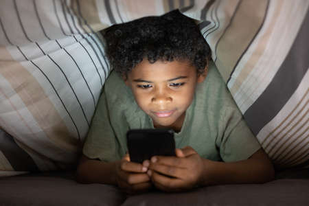 Content cute Black boy with curly hair lying on bed and using smartphone under duvet at nightの写真素材