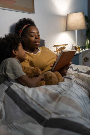 Careful African American blackwoman lying under duvet in bed and reading fairy tale to sonの写真素材