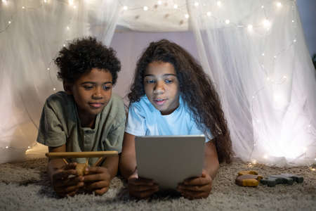 Elder African American sister with wavy hair lying on carpet and reading book on tablet to brother inside of play tentの写真素材