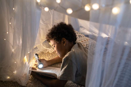 Curious Black boy with curly hair lying inside of play tent decorated with Christmas lights and...