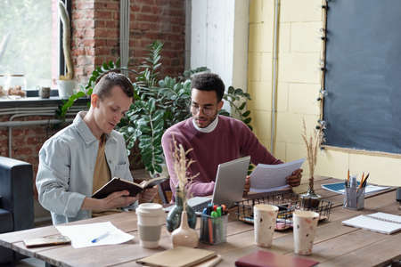 Two young intercultural businessmen in casualwear planning work while one of them looking at his notes in notebookの写真素材
