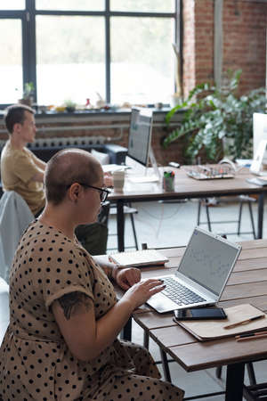 Contemporary young businesswoman sitting by table in front of laptop while networking against male colleagueの写真素材