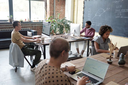 Group of young contemporary economists working with financial data while sitting by tables in front of computers in officeの写真素材