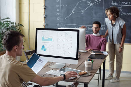 Young economist looking at financial data on computer screen while sitting in front of colleagues...