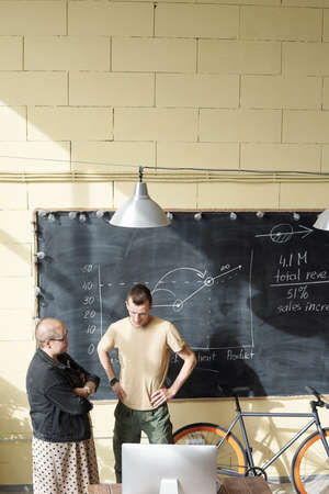 Two young economists standing by blackboard in front of computer monitor and discussing online data...