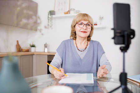 Blond mature woman in eyeglasses speaking in front of smartphone camera during livestream or online trainingの写真素材
