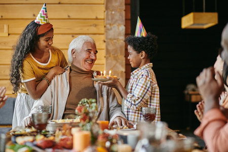 Happy aged man looking at his interracial grandson with birthday cake while sitting by festive table...
