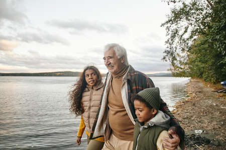 Cute biracial girl looking at her grandfather and listening to him during walk along waterside on...