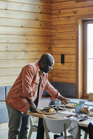 Young African man putting homemade cooked food on festive table prepared for family dinner in living-roomの写真素材