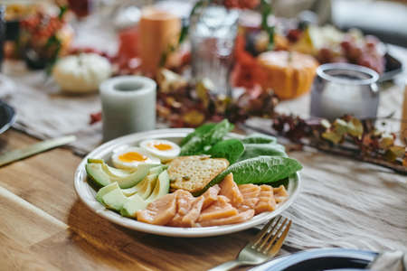 Plate with sliced fresh avocado, leaves of spinach, halved boiled egg, cookie and chopped fish standing on served wooden tableの写真素材