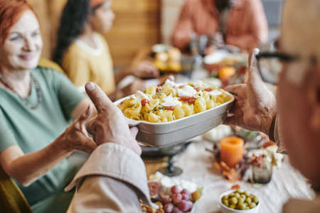 Hand of mature man passing bowl with baked pasta to his wife while both sitting by festive table against their familyの写真素材