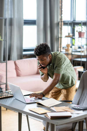 Serious young Blackman standing at table and checking information on laptop while talking by phone in officeの写真素材